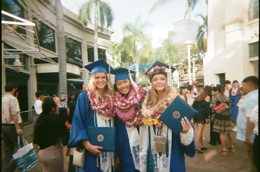 Graduates in blue gowns and leis holding diploma covers during an outdoor campus graduation ceremony.