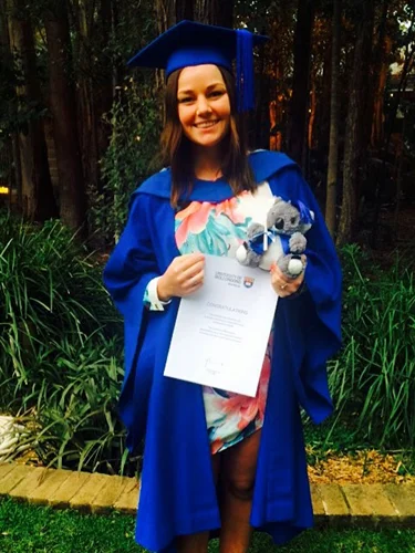 Graduate in blue gown holding a certificate while standing outdoors among trees.
