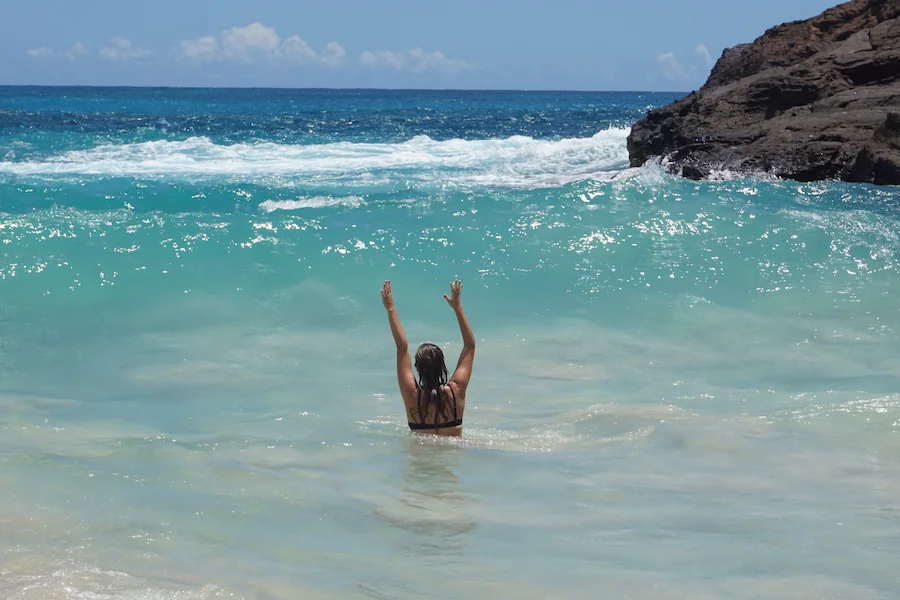 Person standing in turquoise ocean water with raised arms, facing waves near a rocky coastline.