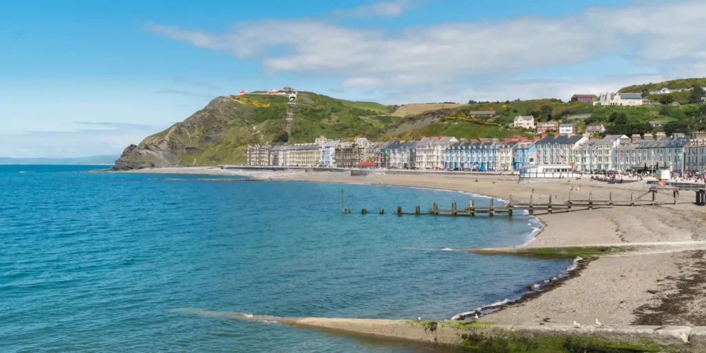 Strandpromenaden i Aberystwyth med fargerike hus, sandstrand og grønn åsside i Wales.
