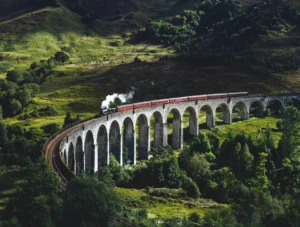 A steam train with red passenger carriages crosses a long stone viaduct through rolling green hills and dense woodland, viewed from above in dramatic sunlight and shadow.