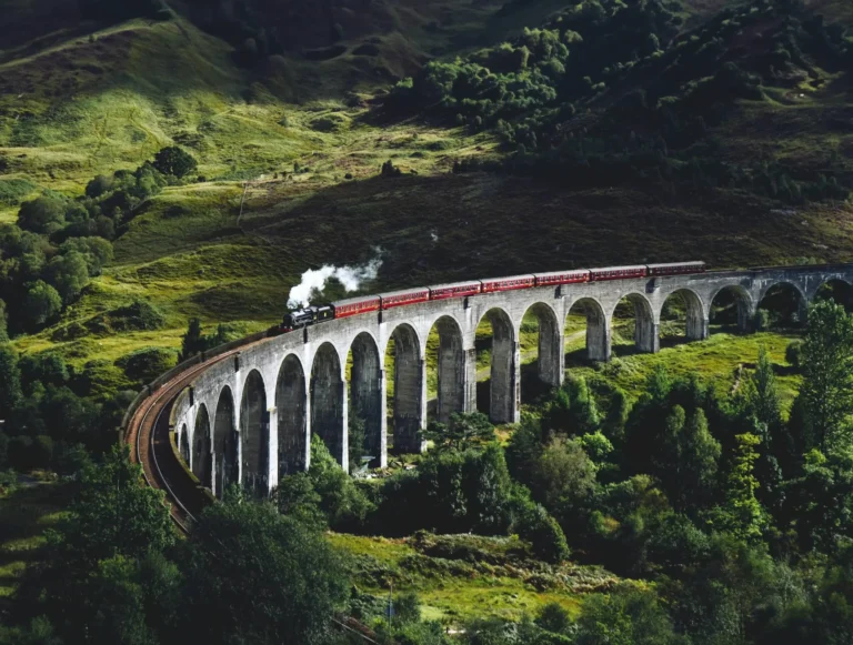 A steam train with red passenger carriages crosses a long stone viaduct through rolling green hills and dense woodland, viewed from above in dramatic sunlight and shadow.