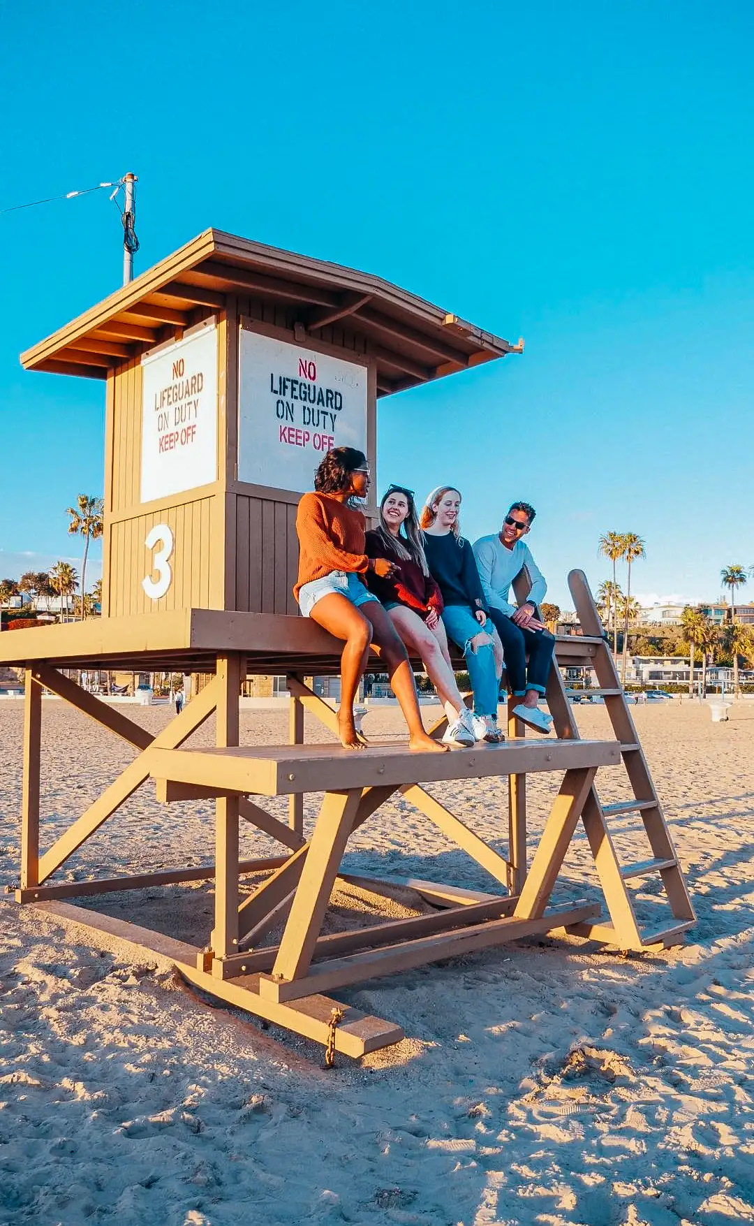 Fire utvekslingsstudenter sitter på et livredertårn på stranden i California, ler og koser seg i ettermiddagssolen. Palmetræer og knallblå himmel i bakgrunnen. Bildet fanger akkurat den følelsen av frihet og fellesskap som mange husker best fra studietiden i utlandet.